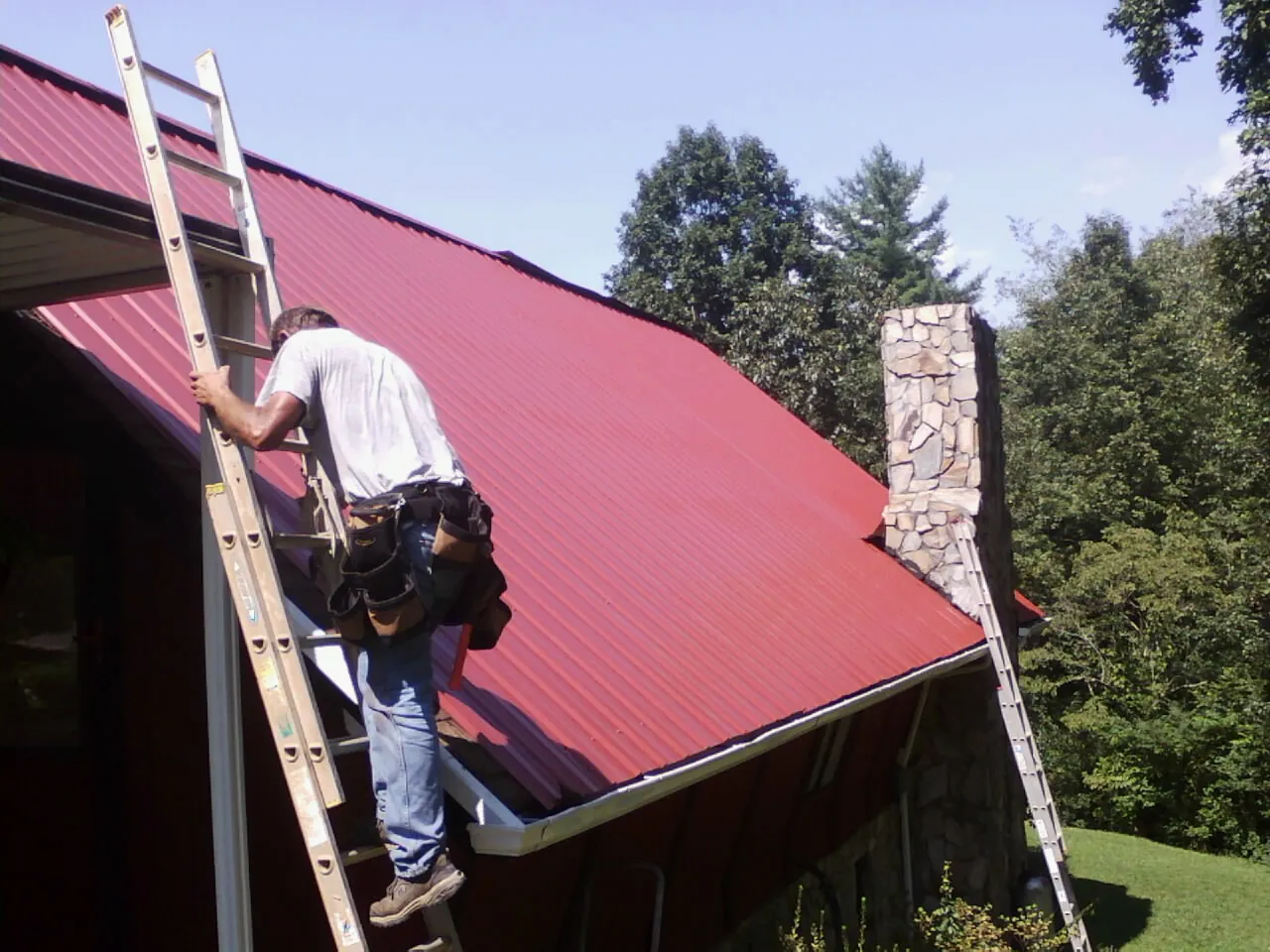 Roofer on a ladder beside a red metal roof and stone chimney — Soffit & Fascia Repair by Roofing Brothers in Olean